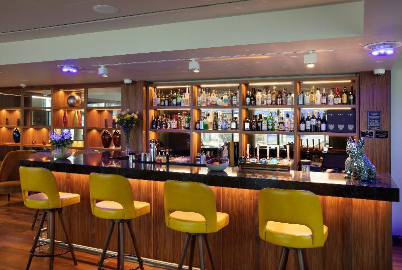 Tower Suites By Blue Orchid London Exteriör bild The photo shows a modern bar area. In the foreground, there are several yellow bar stools lined up in front of a sleek black countertop. Behind the bar, there is a well-stocked shelf displaying a variety of bottles, including spirits and liqueurs, il