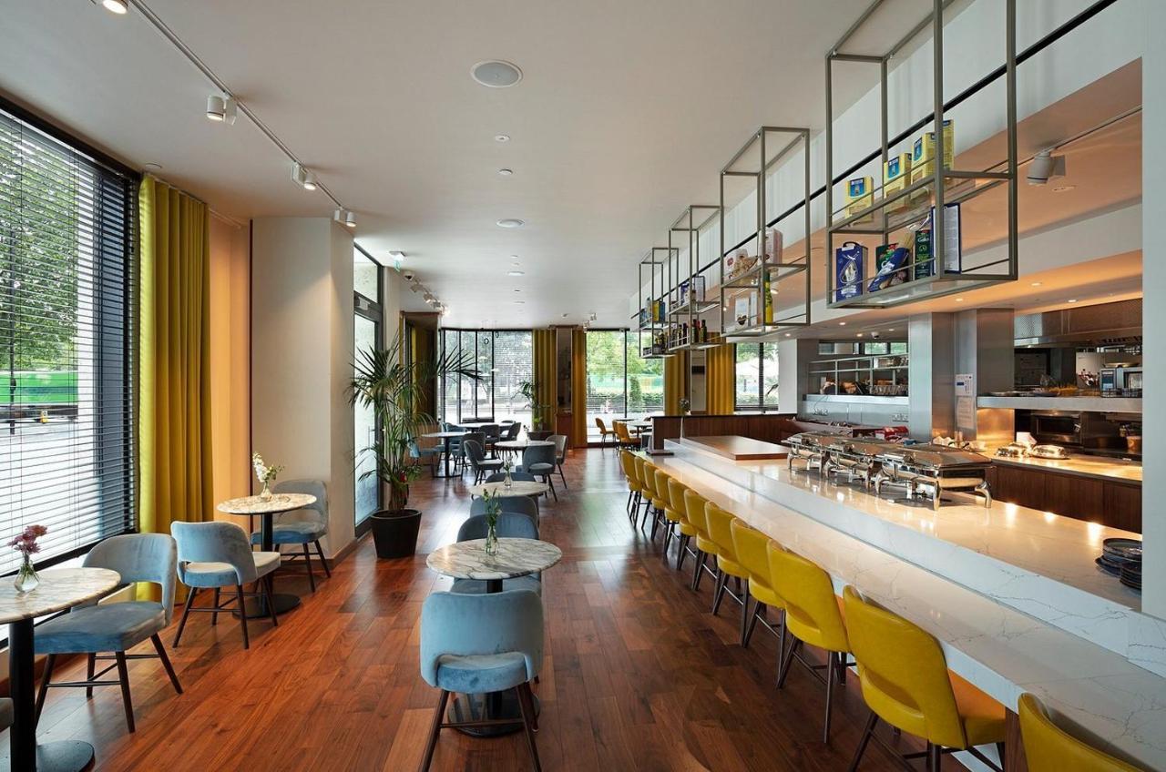 Tower Suites By Blue Orchid London Exteriör bild The photo shows a modern restaurant interior. It features a spacious layout with large windows allowing natural light to flood the space. The decor includes a combination of wooden flooring and colorful furniture—blue and yellow chairs arranged aroun