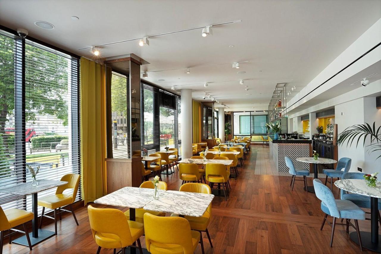 Tower Suites By Blue Orchid London Exteriör bild The photo depicts a modern dining area with a bright and airy atmosphere. There are several tables with marble tops, accompanied by yellow chairs, creating a vibrant contrast with the wood flooring. The space features large windows that allow ample n