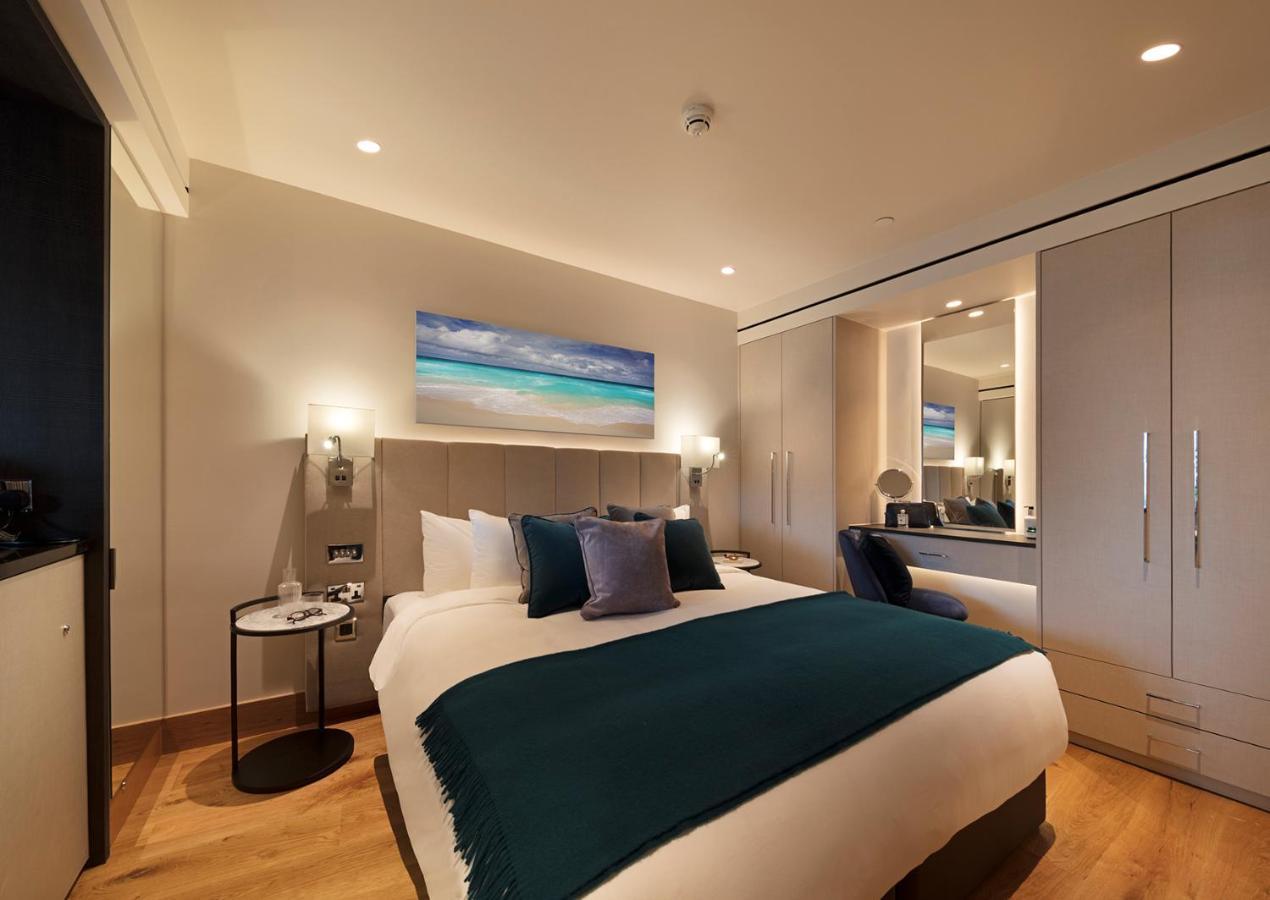 Tower Suites By Blue Orchid London Exteriör bild The photo shows a modern hotel room featuring a double bed with a white duvet and a dark teal throw blanket. There are decorative pillows in shades of blue and purple on the bed. To one side, there's a round bedside table with a small lamp. A large m