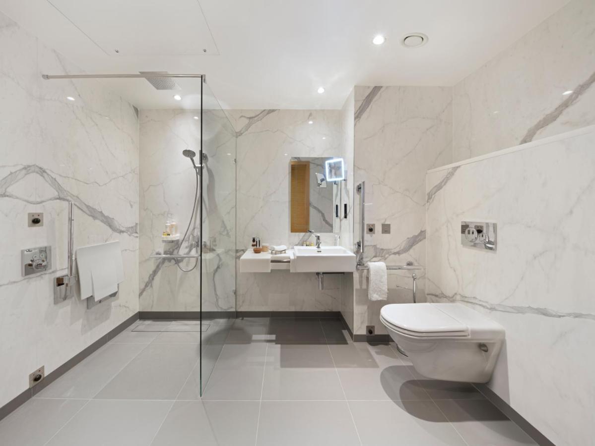 Tower Suites By Blue Orchid London Exteriör bild The photo shows a modern bathroom that features marble walls with a light color. There is a large walk-in shower with a glass partition, and the shower area has a rain shower head. To the right, there is a stylish sink with a mirror above it, and var