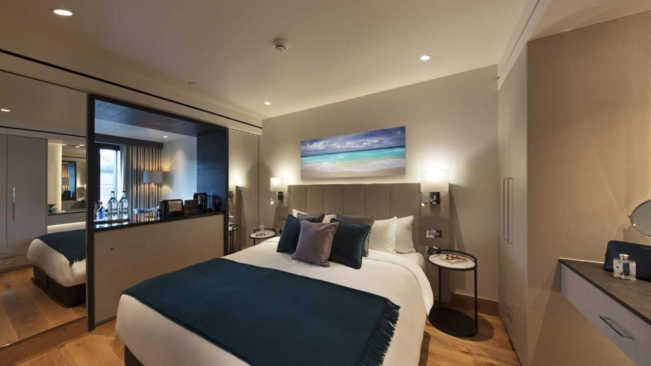 Tower Suites By Blue Orchid London Exteriör bild The photo shows a modern hotel bedroom featuring a large bed with a light-colored headboard and a deep blue throw at the foot. The bed is adorned with multiple decorative pillows. On the wall above the bed, there is a colorful painting depicting a be