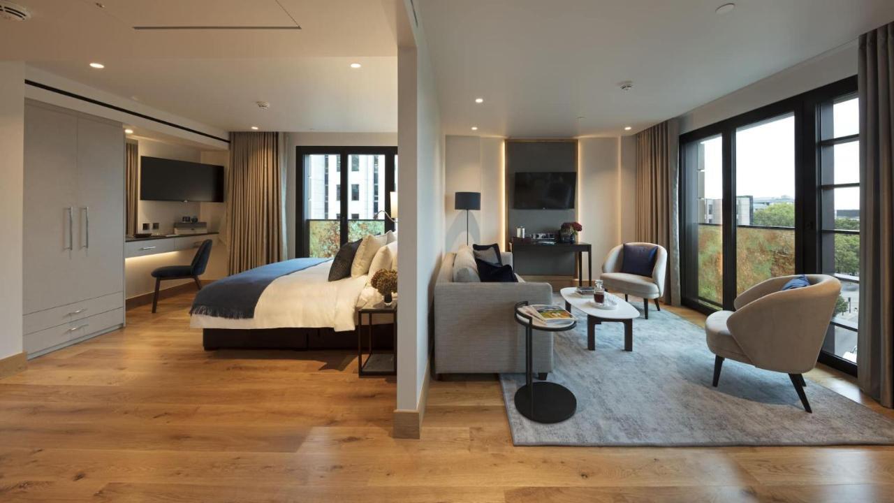 Tower Suites By Blue Orchid London Exteriör bild The photo shows a modern hotel room or suite. On one side, there's a bed with a blue and white duvet and pillows, accompanied by a bedside table with a lamp. Opposite the bed, there's a living area featuring a cozy gray sofa, a small round coffee tab