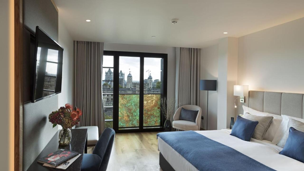 Tower Suites By Blue Orchid London Exteriör bild The photo shows a modern hotel room featuring a large bed with blue and white bedding, a cozy armchair, and a small desk. There is a flat-screen television mounted on the wall across from the bed. The room has large glass doors leading to a balcony o