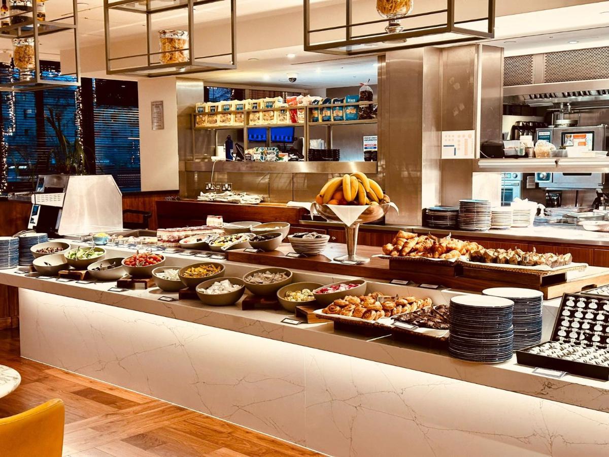 Tower Suites By Blue Orchid London Exteriör bild The photo depicts a buffet-style setup, likely in a restaurant or a hotel dining area. In the foreground, there are multiple bowls and platters filled with a variety of food items, including salads, fruits, pastries, and other dishes. There is a long